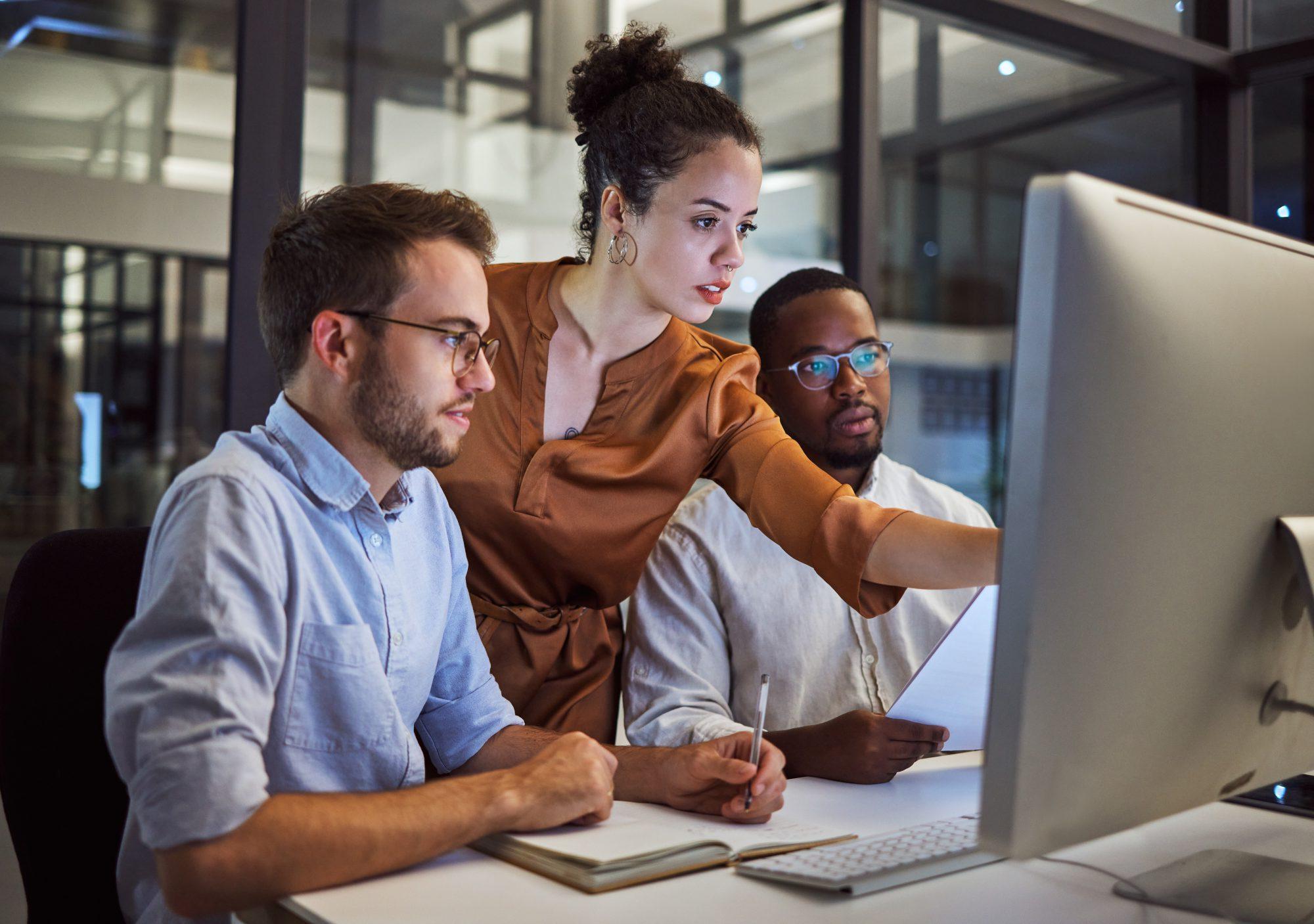 Three individual in front of a computer. One of them is pointing to the screen while the other two appear to be listening.